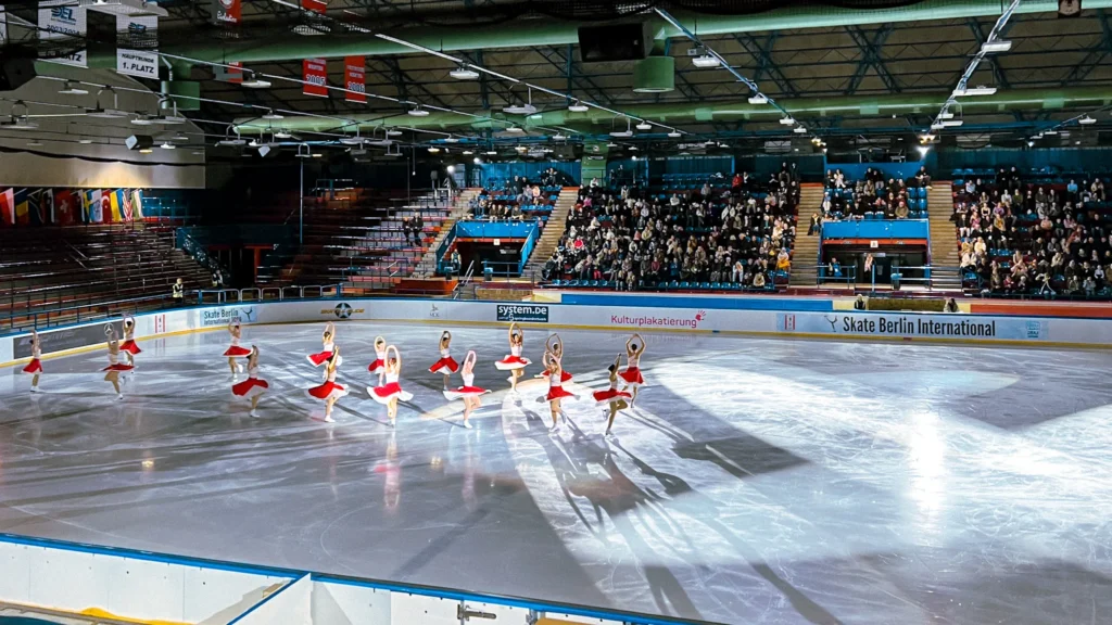 Photo: Florian Knorn A group of skaters in red skirts on the ice of the “Wellblechpalast” at SBI26, with full stands behind.